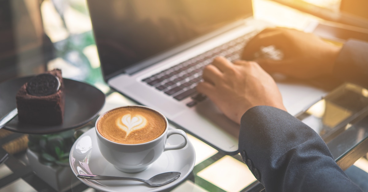 Hands typing on a MacBook next to a cup of coffee with a foam heart