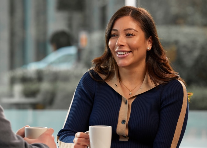 Woman with coffee cup sits down to talk with a male coworker.
